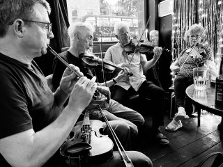 Three fiddlers and a tin whistle player playing in a semi-circle at a pub table.