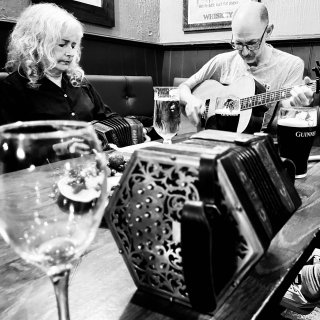 A concertina and a wine glass sit in a table. On their side a woman is playing concertina accompanied by a man on guitar.