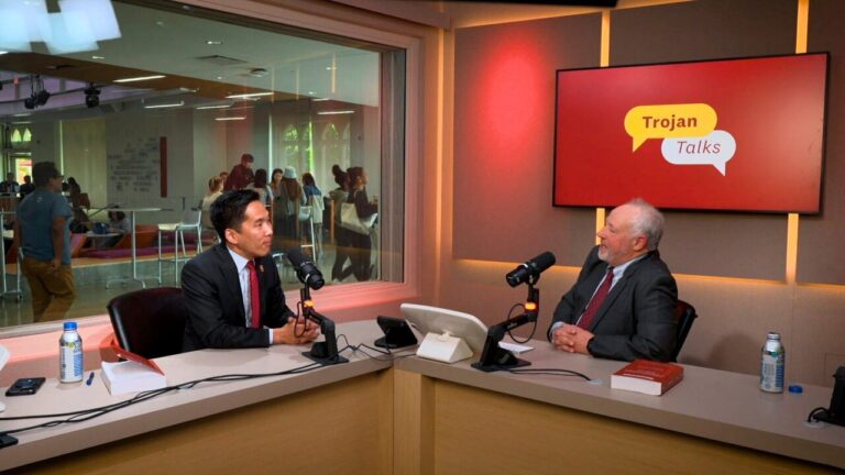 Two men sit in sound booth and speak into microphones.