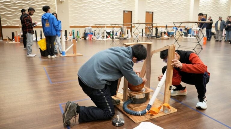 Two young men assemble a contraption of wood, tubing and a basketball as others do similar