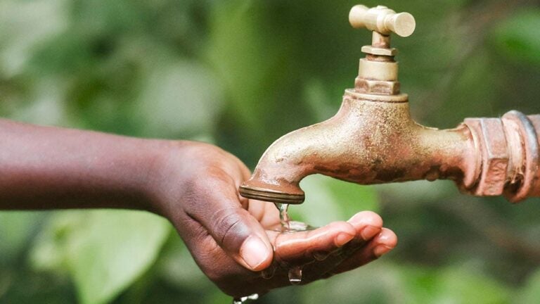 Close-up of a hand under a brass water tap as water drips into the palm, with a blurred green background.