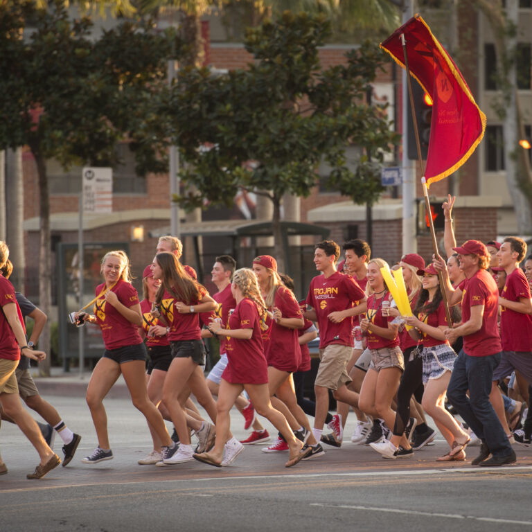 Group of USC students run to the Residential College Cup holding the flag of their Residential flag and wearing the matching t-shirts.