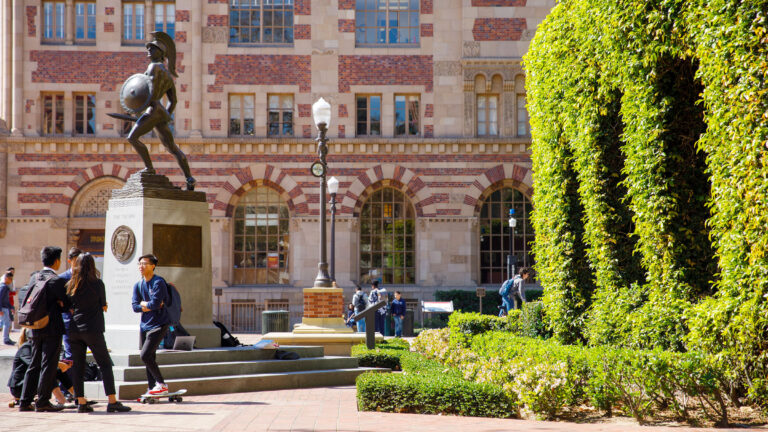 A side view of the statue, Tommy Trojan, next to a large bush with five students gather around the base chatting.