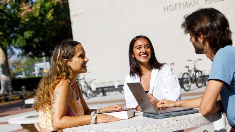 Three students sit on a stone table outdoors and talk with their study materials around them.