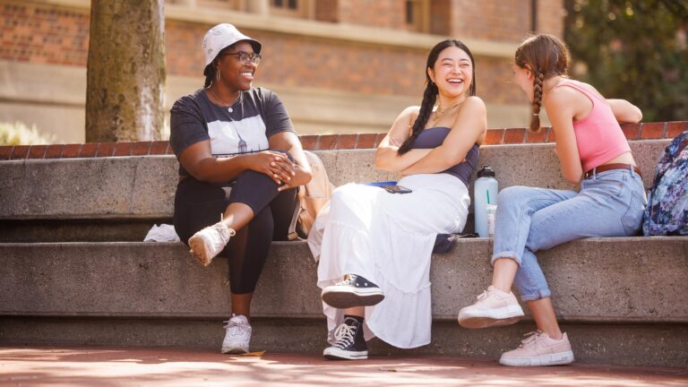 Three students sitting on a bench laughing and talking on a bright sunny day.