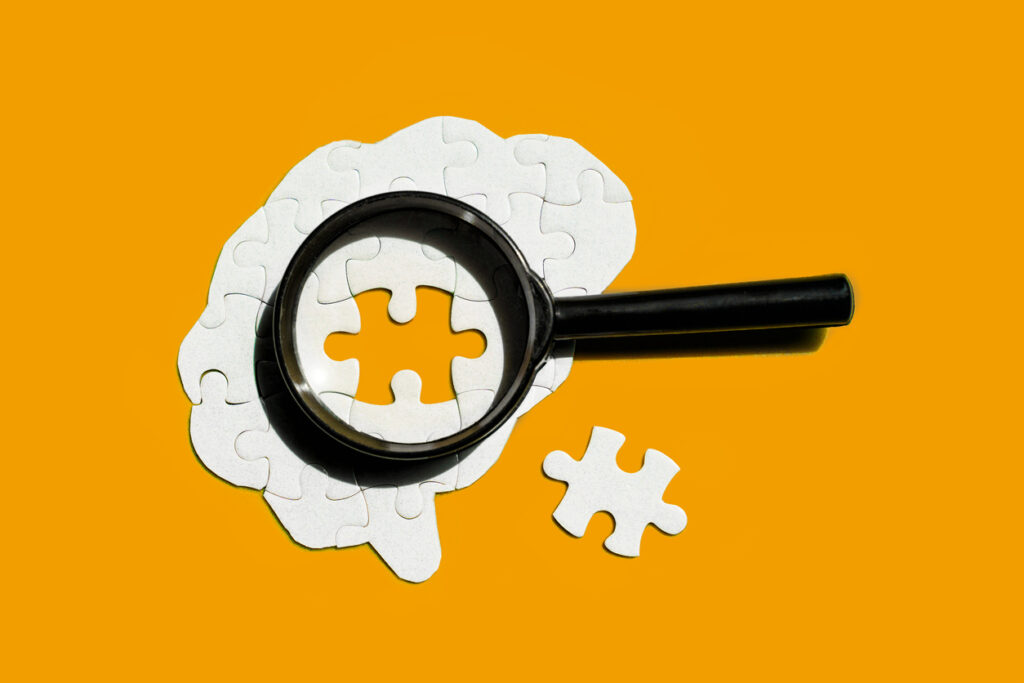 Brain made of white puzzle pieces, with a magnifying glass focusing on an empty puzzle piece isolated on yellow backdrop. Mental health, cognition, and psychological well-being concept.