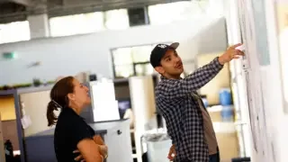 Two students discussing their work in front of a board inside a building.