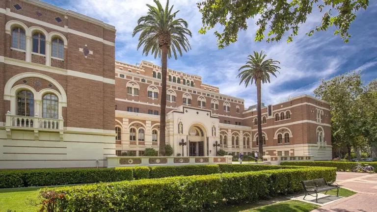 Two large palm trees stand in front of the USC Doheny Library against a bright blue sky on a sunny day.