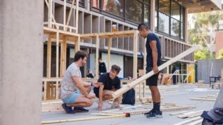 Three students talk together as they build a wooden structure and other partially built structures stand in the back at the USC School of Architecture.