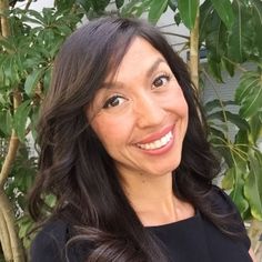 a woman with long dark hair smiling at the camera and wearing a black top in front of a tree