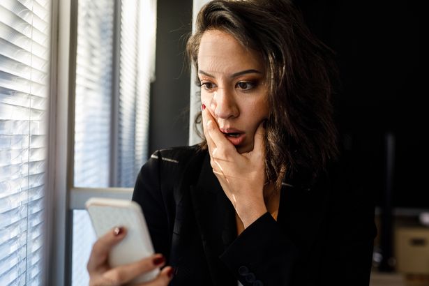 Portrait of shocked young woman standing by the window, hand over mouth, reading a worrying text message via smart phone.