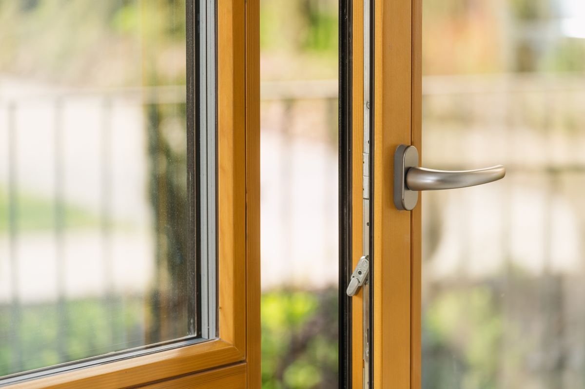 Close-up of a modern wooden window with a silver handle, open slightly. Balcony door in apartment. Blurred summer backyard against clear transparent glass, scenic view from room