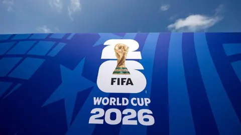 A Photo of the FIFA World Cup 2026 logo in white letters which features a gold soccer trophy with emerald green accents near the bottom against a blue flag backdrop and a baby blue sky. 
