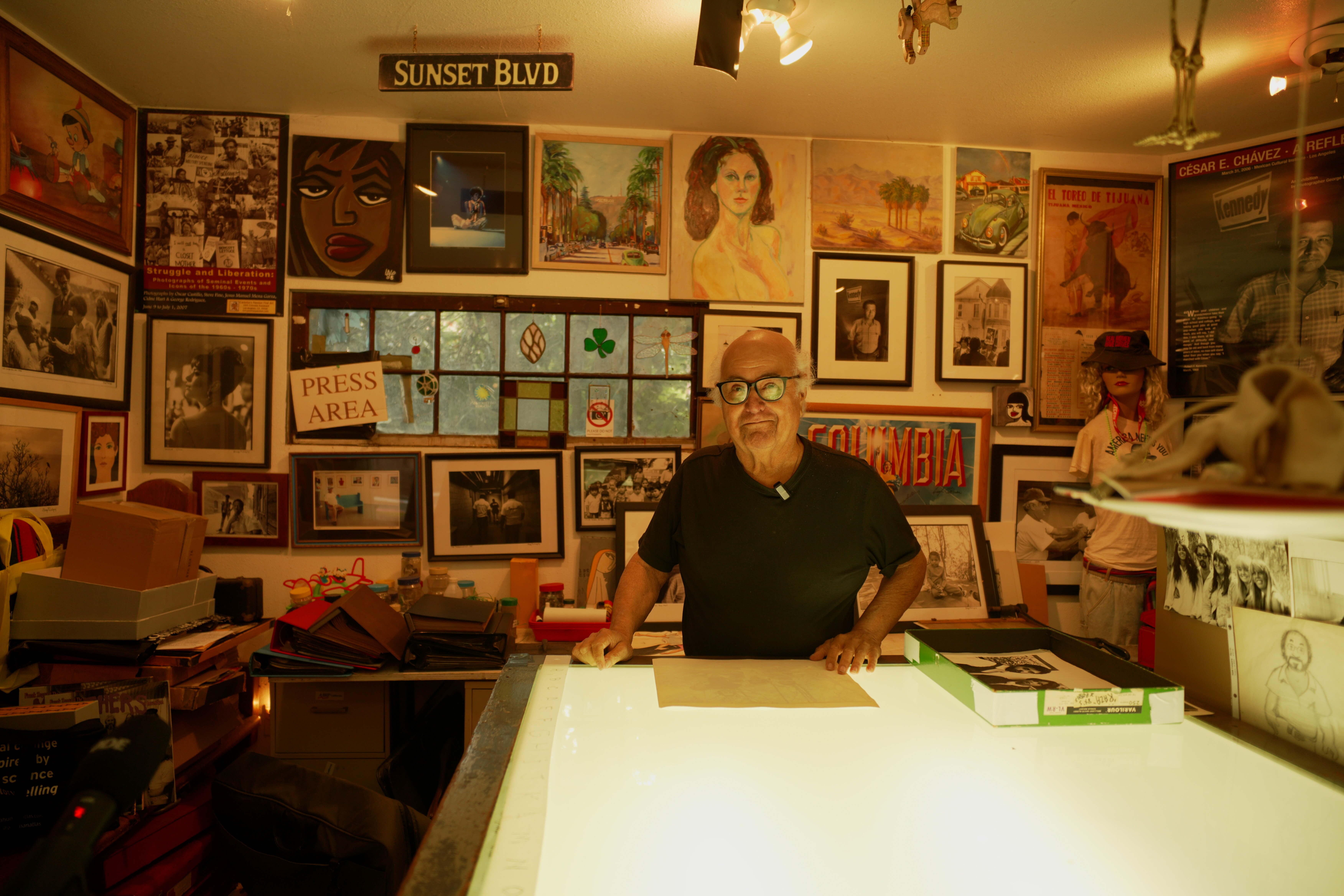 George Rodriguez leans against a light table in front of a gallery wall full of art, photography, and prints.