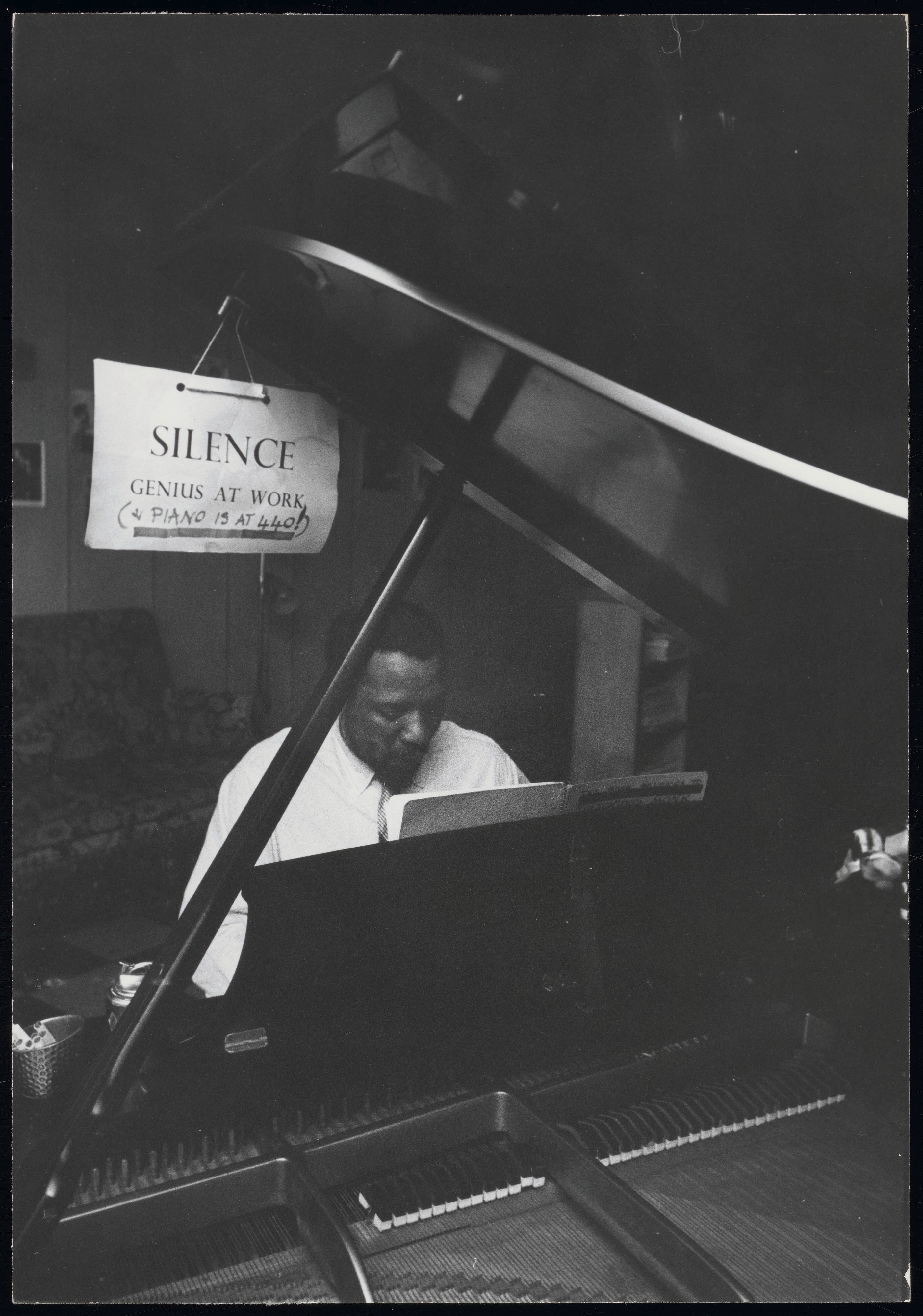 Thelonius Monk sits at a piano under a sign reading "SILENCE Genius At Work"