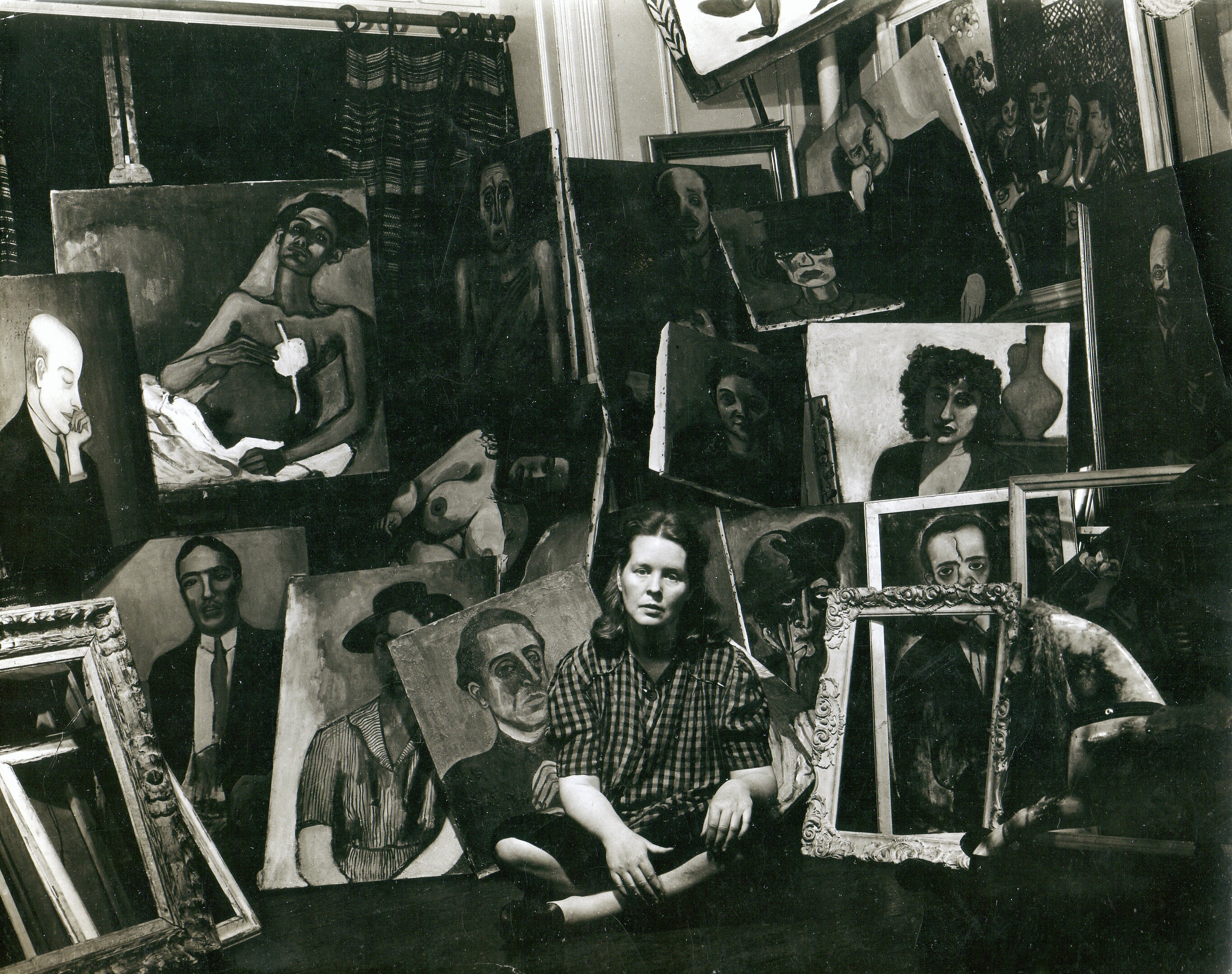 Black and white photograph of artist Alice Neel sitting on the floor amidst a riot of paintings and frames.